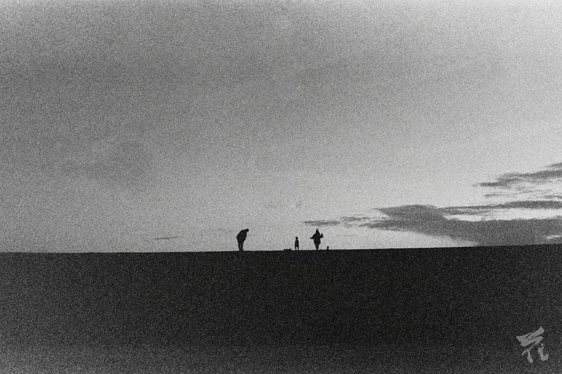 Black and white analog photograph showing three people's silhouettes in the distance. The bottom portion of the picture is black, the top is a backdrop of clouds. One of the persons is inclined forwards with their hands on their knees towards the other two. The second seems to be a child running around. The third a person with their arms open wide.