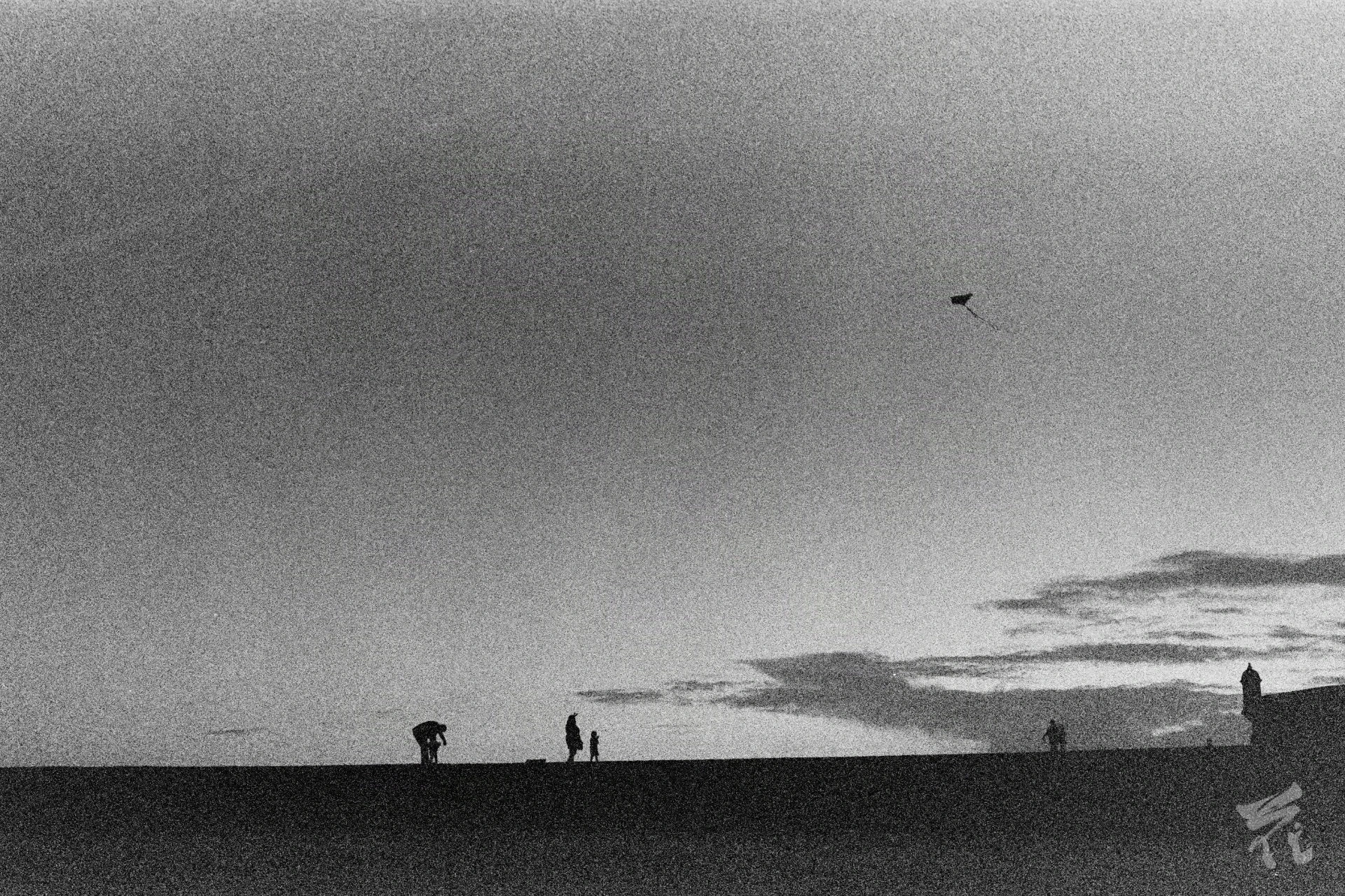 Black and white analog photograph showing four people's and an observation deck's silhouettes in the distance. The bottom portion of the picture is black, the top is a backdrop of clouds. One of the persons is inclined forwards with attending a child, the other two are flying a kite that is visible in the upper right portion of the photograph.