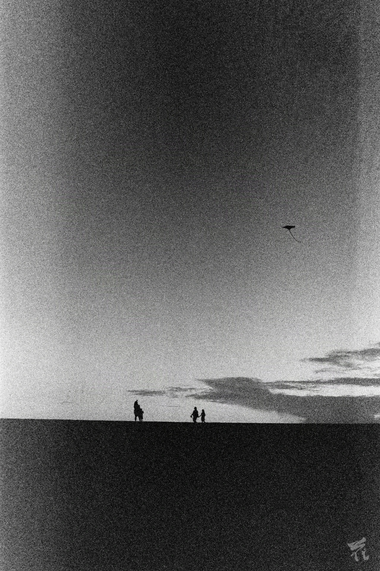 Black and white analog photograph showing four people's and an observation deck's silhouettes in the distance. The bottom portion of the picture is black, the top is a backdrop of clouds. One of the persons is inclined forwards with attending a child, the other two are seemingly walking together.