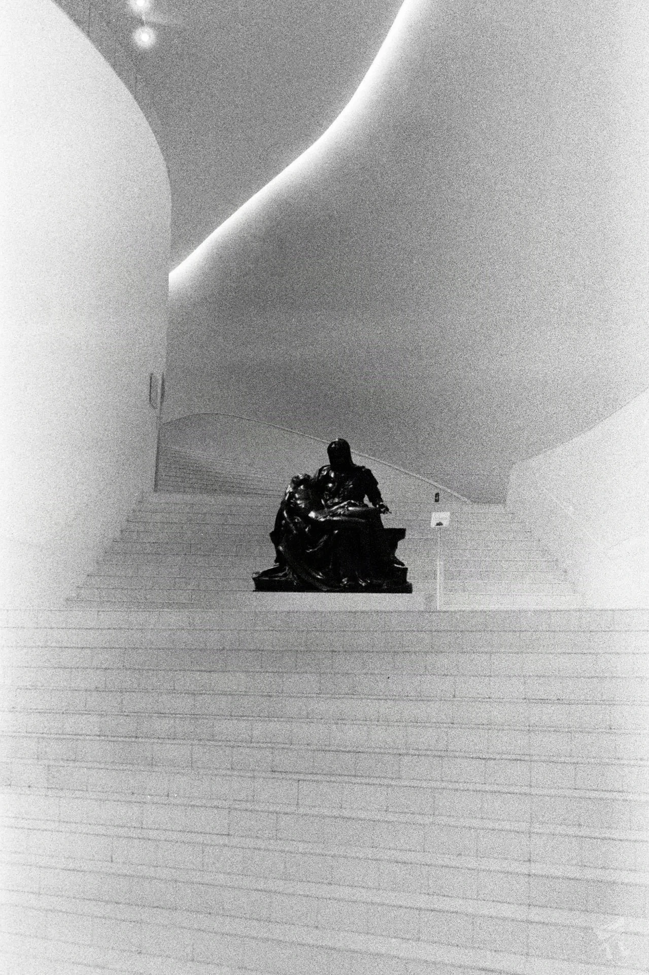 An analog photograph of a very wide white and winding stairwell. The camera is panned upwards. In the center of one of the flat resting zones of the stairs suts a dark black bronze sculpture of a naked male-looking person holding a naked female looking person. They are covered in sheets of cloth draped over them. The woman appears to be curled up in anguish. The white light from the stiars bounces sharply off each person's body.