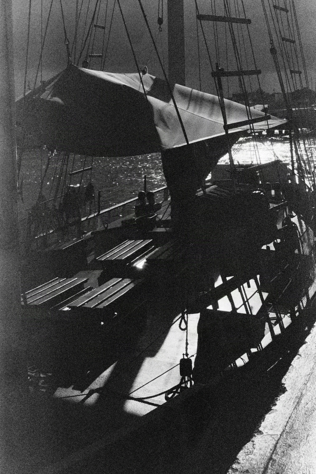 A black and white analog photograph. Stark contrast shows shadows cast by the sun reflecting off the deck of a boat opposite to where the picture was taken. Many sails, wooden pallets and barrels line the deck. In the foreground, ropes go up to the mast.