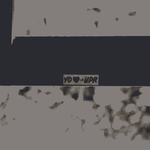A black and white analog photograph showing the top of a wooden beam in the foreground. On the wooden beam, in the center of the image a whitr sticker with the text "YO ❤️ la UPR" (EN: I LOVE THE UPR). The background is far away and out of focus.
